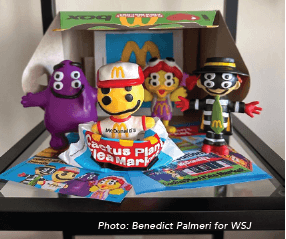 A photo of a McDonald’s Happy Meal box that is opened to show four stylized collectible toys standing in front of a blue mat with cartoon characters on it. From left to right, the toys are a purple Grimace, a yellow McDonald’s character with a red hat, a red-and-yellow-striped Birdie, and a Hamburglar in a black and white striped outfit and a black hat. The McDonald’s and ‘Cactus Plant Flea Market’ logos are visible on the Happy Meal box and the mat. The toys are from a limited-edition collaboration. The bottom of the image has a credit that reads ‘Photo: Benedict Palmeri for WSJ’.