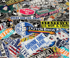 A close-up view of a large collection of colorful stickers, many of which are associated with the Dutch Bros coffee chain. The stickers are layered on top of each other, creating a collage effect. Prominent stickers include a large circular blue and white one with a stylized windmill, a black and yellow sticker that reads ‘CAFFEINE IS NOT A CRIME’, and several blue and white stickers with the phrase ‘DRINK ONE FOR DANE’. There are also stickers with American flag designs, stylized logos, and other phrases. The background appears to be a black-and-white patterned surface. The bottom right corner has a credit that reads ‘Photo: Dutch Bros’.