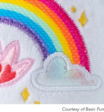 A close-up shot of the embroidered design on a Care Bears stuffed animal’s belly. The design features a colorful rainbow arching over a silver cloud. A pink, flame-like emblem is partially visible to the left, and a couple of yellow embroidered stars are scattered around the rainbow and cloud. The fabric is a soft, white plush material. The bottom right corner has a caption that reads ‘Courtesy of Basic Fun’.
