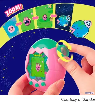 A digital photo of two hands holding a pink and green Tamagotchi device shaped like an egg. The device has a digital screen showing a pink rabbit-like creature in a green field. A finger is turning the green knob at the top of the device, with a yellow arrow showing the direction of rotation. Above the device, a curved strip shows a ‘ZOOM!’ feature with several cartoon images of the pink rabbit and other characters. The background is a starry night sky with a yellow crescent shape. The bottom right corner has a caption that reads ‘Courtesy of Bandai’.