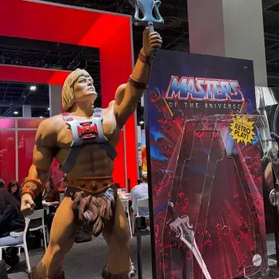 A large, life-sized statue of the He-Man character from the Masters of the Universe franchise. He is a muscular man with blonde hair, wearing his classic harness and loincloth, and is holding his Power Sword up in the air as if he is about to shout ‘By the power of Grayskull!’. Next to him is a large display board for the ‘Masters of the Universe’ toys, which has a large plastic blister pack on the front, similar to old-style toy packaging. The background is a convention hall with red and black booth dividers and other people.