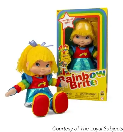 A product shot of a Rainbow Brite doll and its packaging. The doll is a white female character with long yellow hair tied with a blue ribbon, rosy cheeks with a blue star, and a colorful outfit consisting of a red and blue top with a yellow star and a metallic blue skirt. She is sitting with her legs outstretched. Behind her is her packaging, a yellow box with a transparent front displaying the same doll. The box is adorned with a rainbow arching around the title ‘Rainbow Brite’. The bottom right corner has a caption that reads ‘Courtesy of The Loyal Subjects’.