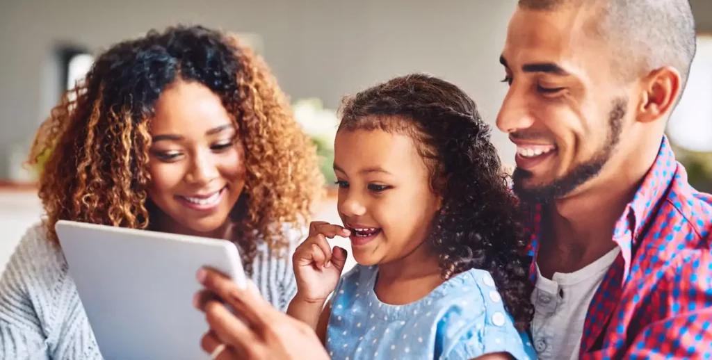 A happy, diverse family of three is gathered together, looking at a tablet. A woman with curly brown hair and a man with a beard are on either side of a young girl with curly hair. The girl is smiling and has a thoughtful expression as she looks at the tablet screen. The man is smiling warmly as he looks at his daughter. The image is bright and appears to be a candid shot of a family bonding.