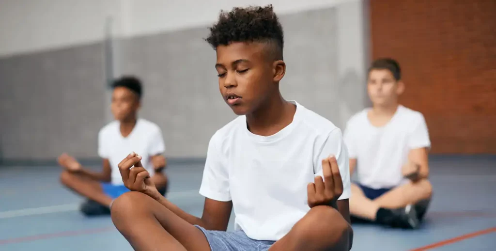 A group of young boys is sitting cross-legged on a gymnasium floor, meditating. The boy in the foreground is a young Black boy with a mohawk haircut. He is wearing a plain white t-shirt and grey shorts, with his eyes closed and his hands resting on his knees, with his thumb and pointer finger touching. There are two other boys in the background, also in white t-shirts and cross-legged, who are slightly out of focus. The floor has light blue and red lines, and the background has a grey wall and a brick wall.