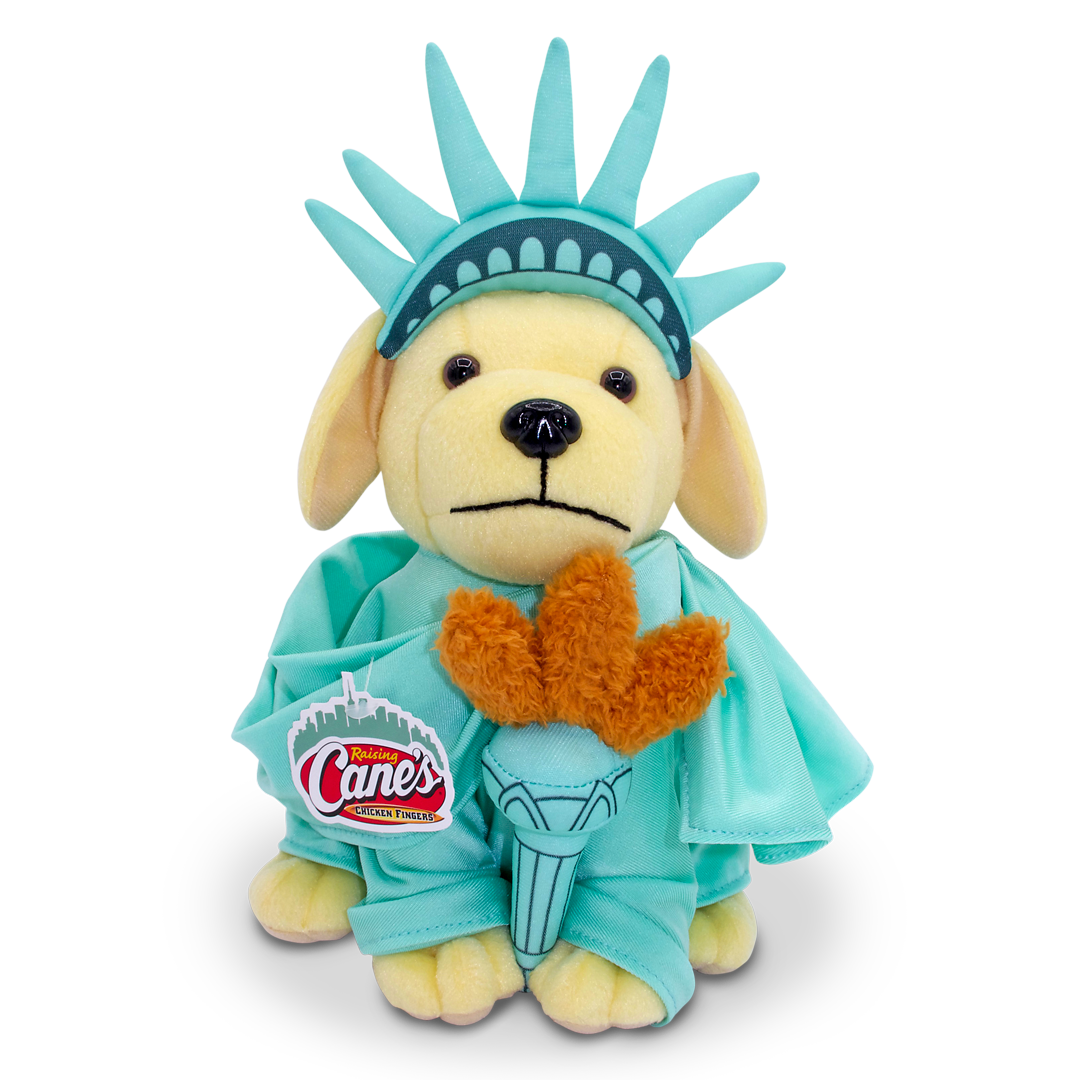 A Raising Cane’s plush puppy dressed as the Statue of Liberty. The puppy wears a teal robe, crown headpiece, and holds a plush orange torch with the Raising Cane’s logo tag on the outfit.