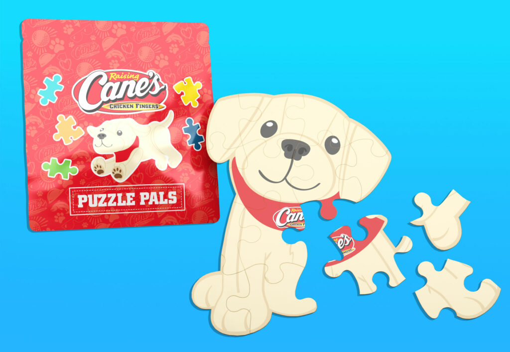 Raising Cane’s “Puzzle Pals” set with packaging and puzzle pieces displayed. The packaging is red with playful graphics and the Cane’s logo. The puzzle depicts the Cane’s puppy mascot with a big smile and red bandana, shown with several detached pieces on a blue background.