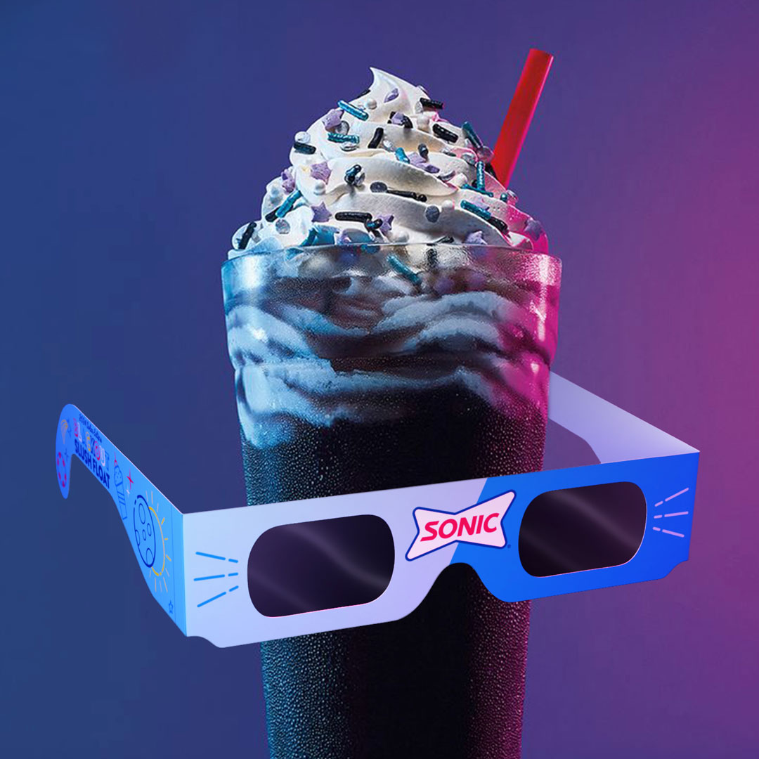 A promotional photo of Sonic’s Blackout Slush Float topped with whipped cream, sprinkles, and a red straw. The drink is paired with branded blue eclipse glasses wrapped around the cup, shot against a neon purple backdrop.