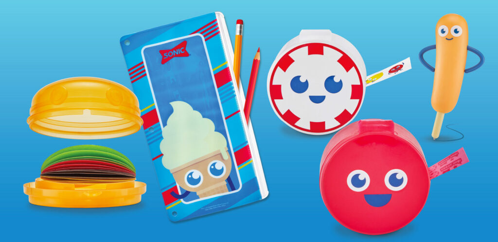 Assorted Sonic-branded kids’ school supplies. Items include a yellow hamburger-shaped case with colorful sticky notes, a blue ice cream cone pencil pouch, peppermint and red button tape dispensers, and a corndog character pen.