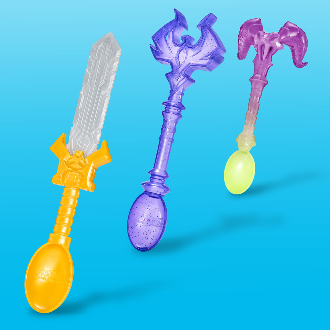 Three plastic collectible spoons inspired by Masters of the Universe, each with fantasy weapon handles. One is orange with a silver sword blade, another is translucent purple with a spiked mace-like top, and the third is translucent purple-pink with a horned ram skull design.