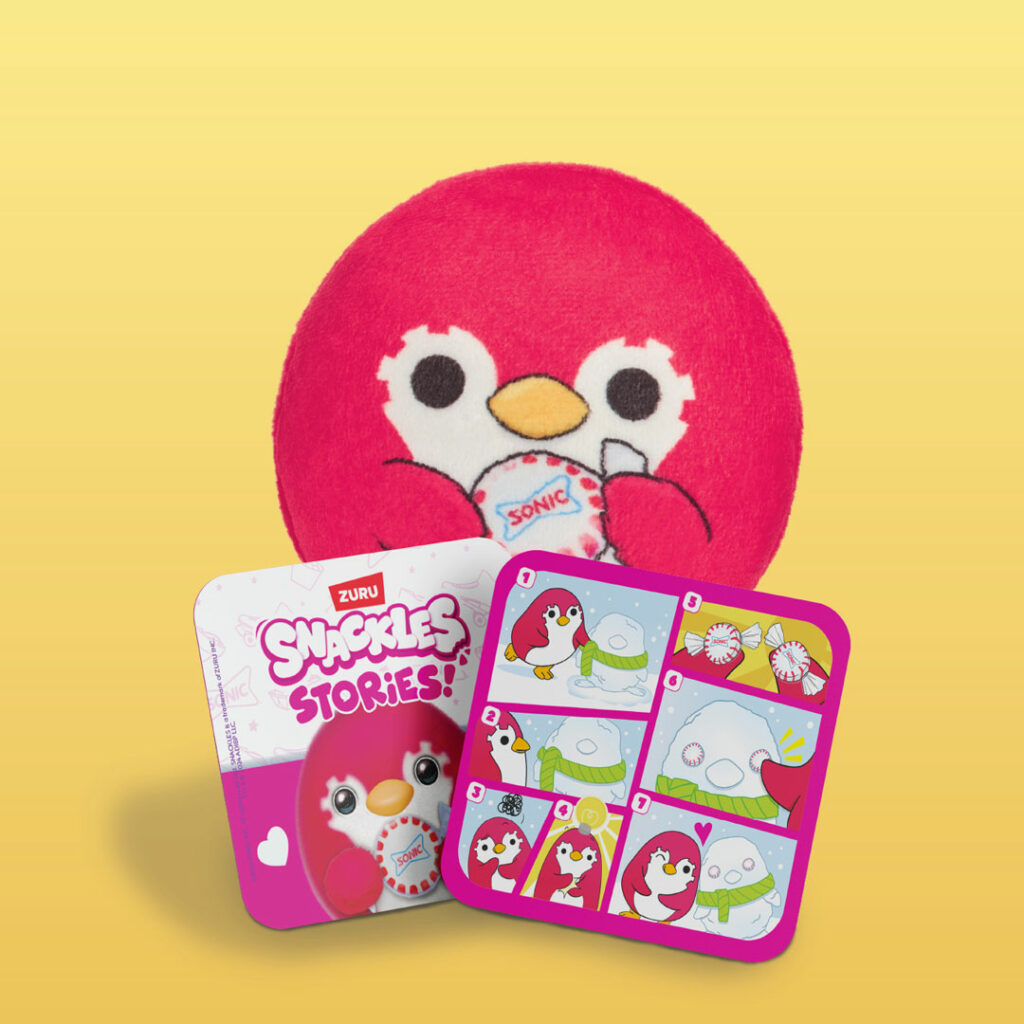 A red peppermint penguin plush toy holding a Sonic cup. In front are two cards: one features the penguin illustration with “Snackles Stories” text, and the other displays a six-panel comic strip of the penguin making a snowman with peppermint candies for eyes.