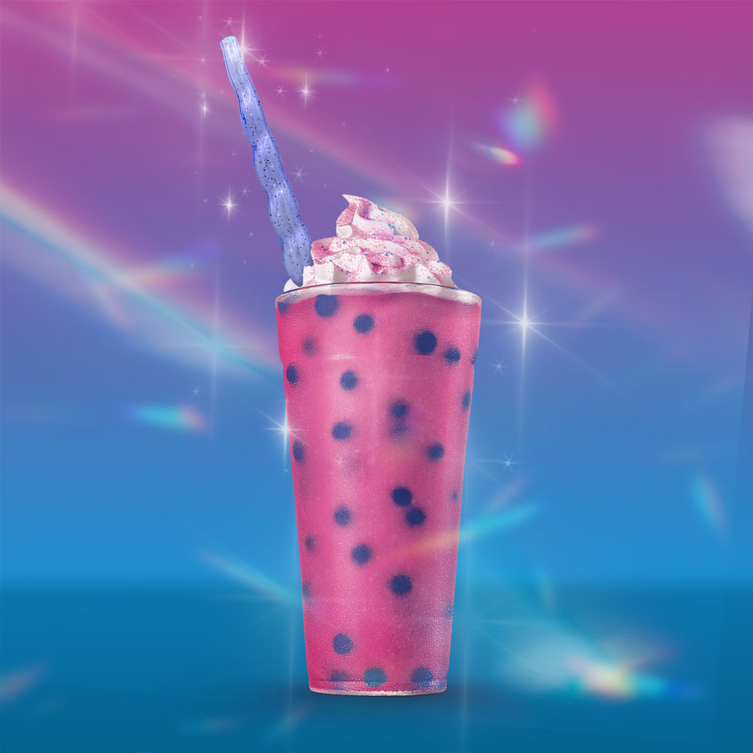 A tall cup of bright pink slush with dark blue popping bubbles is topped with whipped cream and pink sprinkles. A twisted purple unicorn horn straw extends from the drink. The background features sparkles and rainbow light effects.