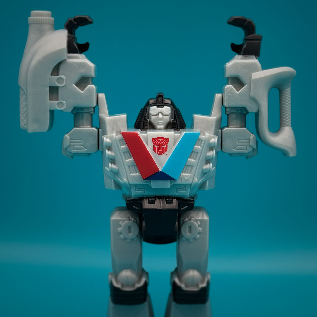 The Valvotron action figure posed outside of packaging against a teal background. The robot has gray and black body parts, raised arms, and the same red and blue V design with the Autobot logo on its chest.