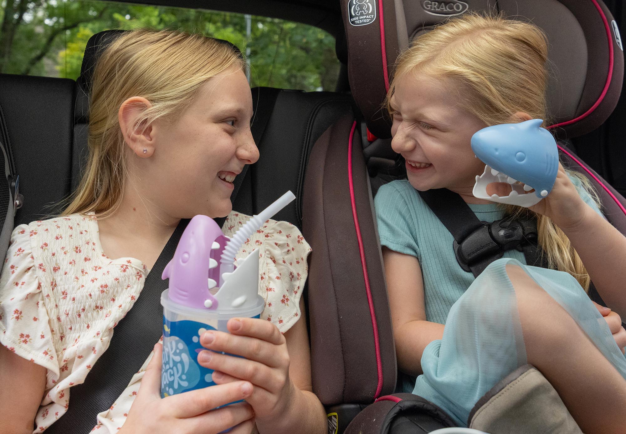 Expertise:Global Production; two gilrs playing with shark puppet cups in the back seat of a car