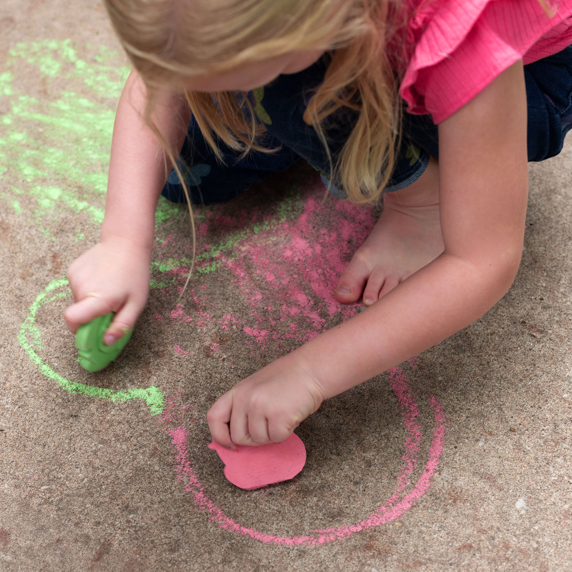 A young girl with sidewalk chalk in each hand, coloring in the shape of a heart on the sidewalk.